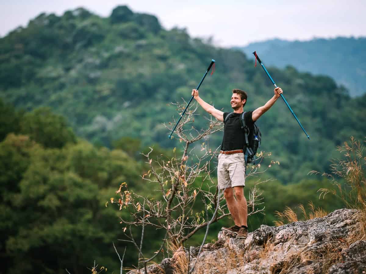 A young man truimphantly raises his walking poles at the top of a tall hill